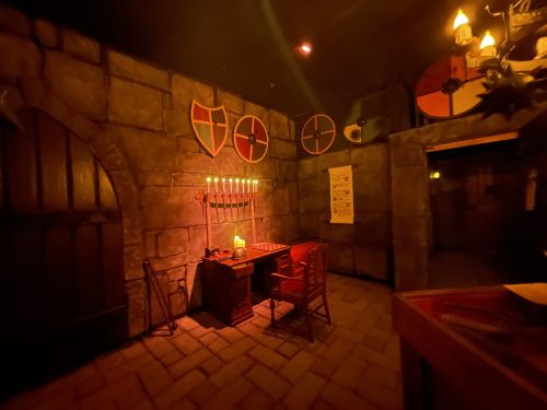 View of a dungeon room featuring a desk, display case, and various shields and weapons on the wall.