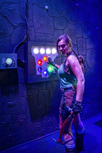 Lara Croft stands before a panel of lit gems of many colors that is wired to a small oscilloscope,